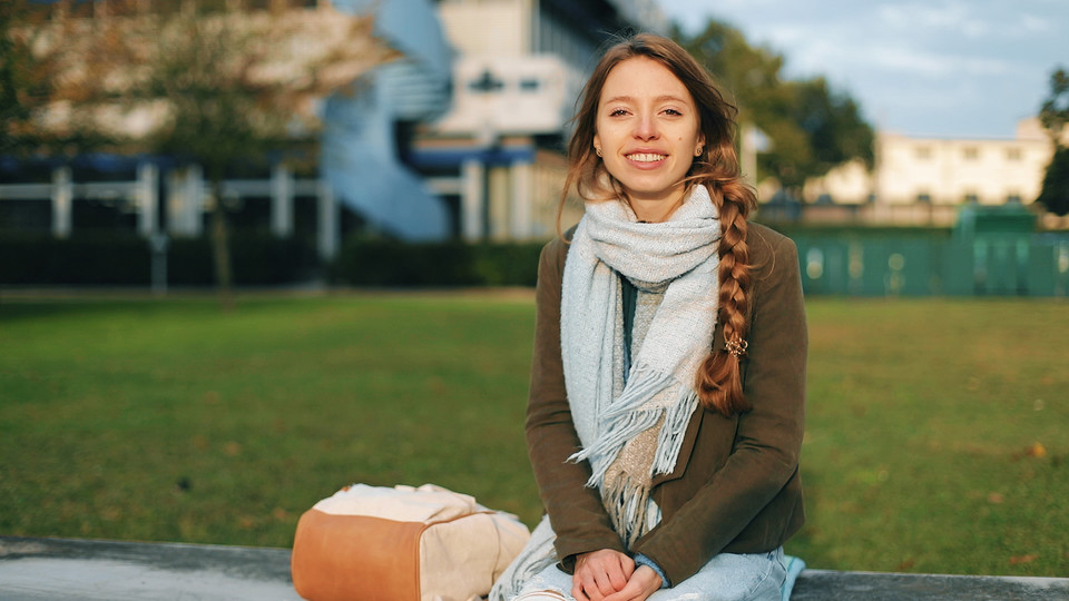 Studentin mit langen braunen Haaren trägt einen geflochtenen Zopf, einen braunen Strickpulli, eine Jeanshose und ein hellblauen Schaal. Sie sitzt vor A5 Bibliothek auf Steinbank und lächelt.